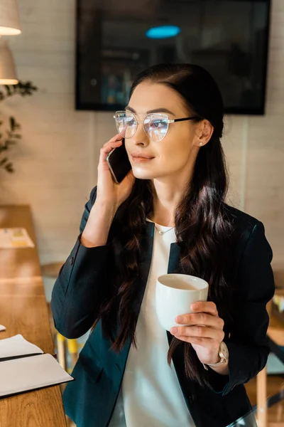 Attraente donna d'affari bruna in occhiali che parla su smartphone e tiene in mano la tazza di caffè in caffetteria — Foto stock