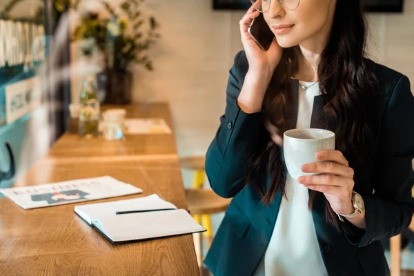 Cropped view of businesswoman talking on smartphone and holding cup of coffee in cafe — Stock Photo