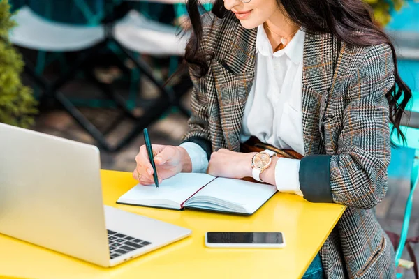 Vista recortada del freelancer femenino que trabaja con planificador, smartphone y laptop en la terraza del restaurante - foto de stock