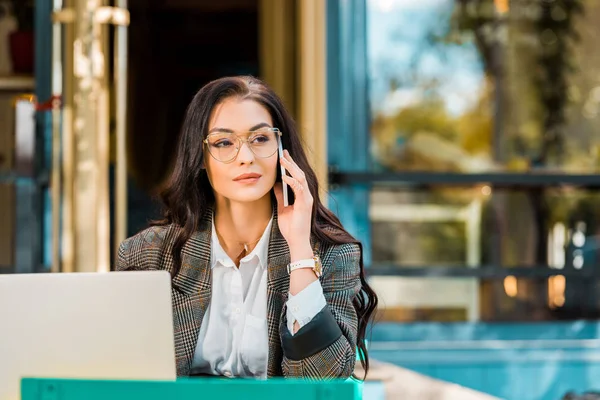 Beautiful freelancer talking on smartphone and working with laptop at restaurant terrace — Stock Photo