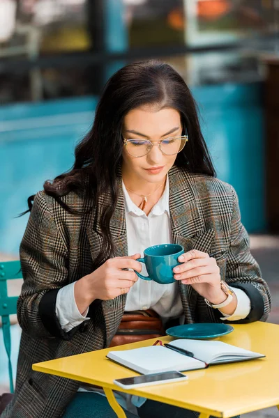 Atractivo freelancer sosteniendo taza de café en la terraza del restaurante con planificador y teléfono inteligente - foto de stock