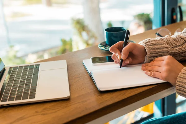 Vista parcial de la escritura freelancer en planificador con teléfono inteligente y portátil en la cafetería - foto de stock