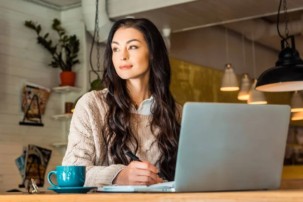Attractive brunette freelancer working with laptop in cafe with coffee cup — Stock Photo