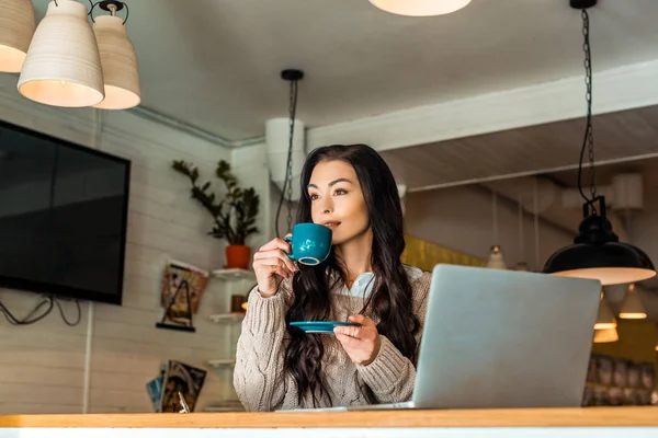 Femme brune en chandail d'automne buvant du café dans un café avec ordinateur portable — Photo de stock