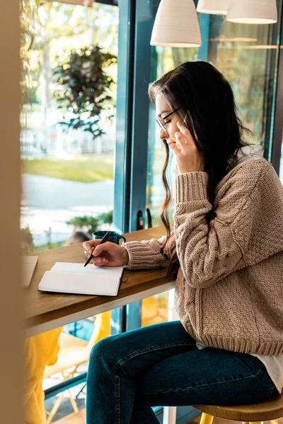 Brunette female freelancer talking on smartphone and writing in diary in cafe — Stock Photo