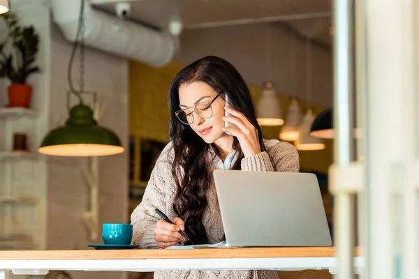 Hermoso teletrabajador hablando en el teléfono inteligente y escribiendo en planificador en la cafetería con el ordenador portátil - foto de stock