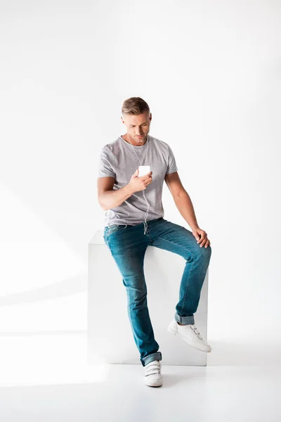 Bell'uomo adulto in t-shirt bianca grigia che ascolta musica con smartphone e auricolari su bianco — Foto stock