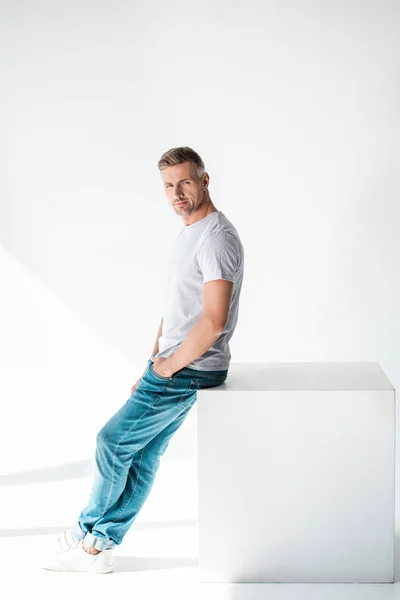 Side view of handsome adult man sitting on cube and looking at camera on white — Stock Photo