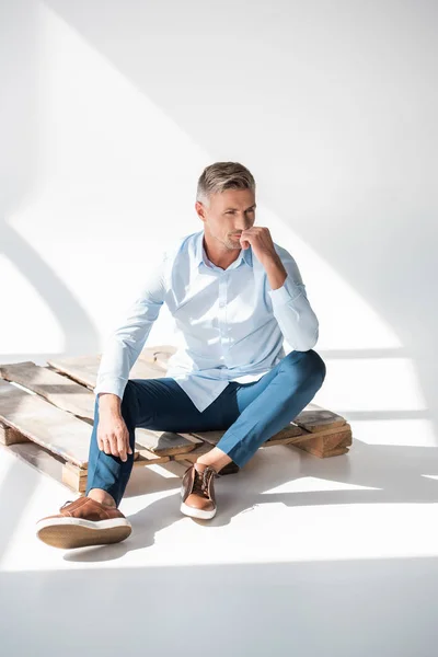 Attrayant homme adulte en vêtements élégants assis sur palette en bois sur blanc et détournant les yeux — Photo de stock