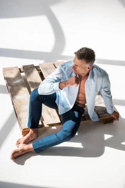 High angle view of attractive adult man sitting on wooden pallet on white and looking away — Stock Photo