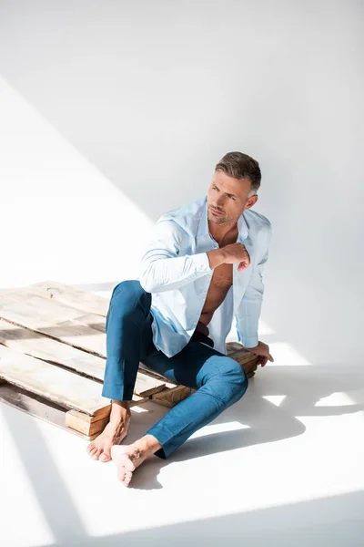 Sexy macho sitting on wooden pallet on white and looking away — Stock Photo