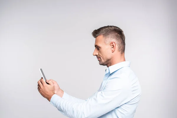 Vista lateral del hombre adulto guapo usando teléfono inteligente aislado en blanco - foto de stock