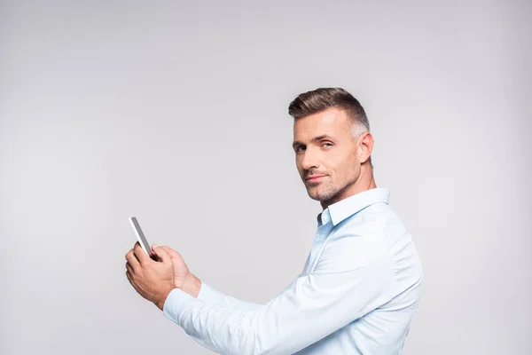 Vista lateral do homem adulto bonito na camisa usando smartphone e olhando para a câmera isolada no branco — Fotografia de Stock