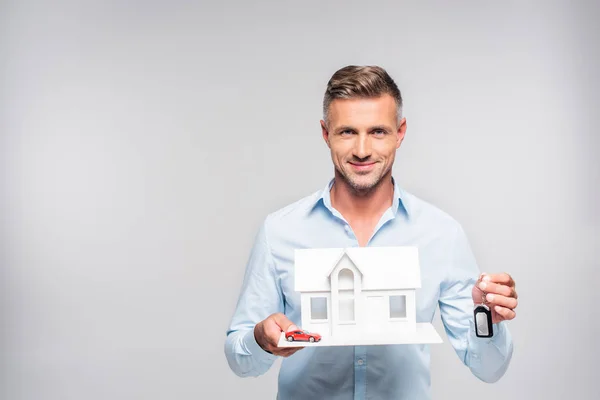 Schöner erwachsener Mann hält Papiermodell eines Hauses mit Autoalarm Fernbedienung und Spielzeugauto isoliert auf weiß — Stockfoto