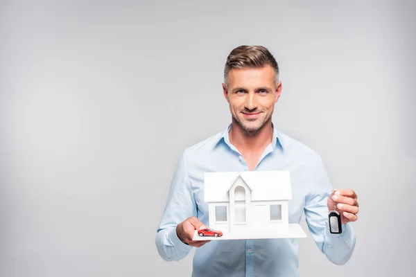 Bonito adulto homem segurando papel modelo de casa com carro alarme remoto e brinquedo carro isolado no branco — Fotografia de Stock