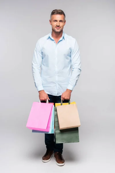 Vista completa de hombre guapo sosteniendo bolsas de compras y mirando a la cámara en gris - foto de stock