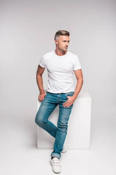 Hombre guapo en camiseta blanca y pantalones de mezclilla sentado y mirando hacia otro lado en gris - foto de stock