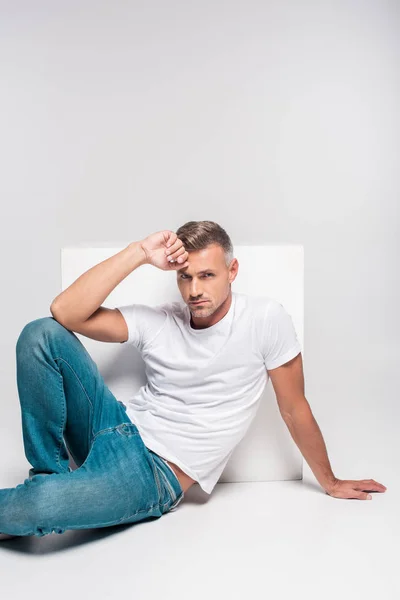 Hombre guapo en jeans y camiseta blanca sentado y mirando a la cámara aislado en gris - foto de stock
