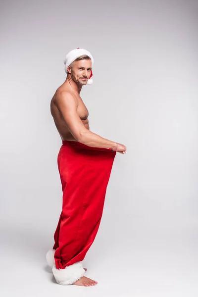 Vue latérale de l'homme torse nu en santa chapeau tenant pantalon rouge et souriant à la caméra sur gris — Photo de stock