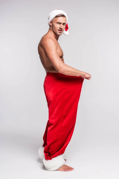 Side view of shirtless man in santa hat holding red pants and looking at camera on grey — Stock Photo