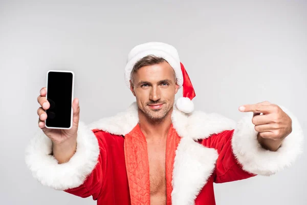 Красивий усміхнений чоловік в костюмі Санти вказує пальцем на смартфон з порожнім екраном ізольовано на сірому — стокове фото