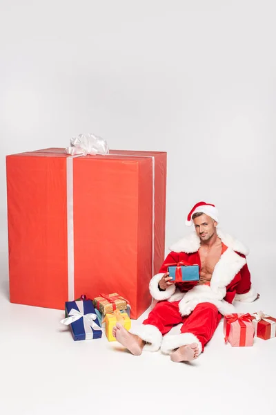 Schöner Mann im Weihnachtsmannkostüm sitzt mit verschiedenen Weihnachtsgeschenken und lächelt in die Kamera auf grau — Stockfoto