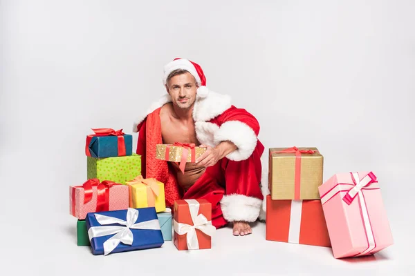 Homme sexy en costume de Père Noël assis avec des boîtes-cadeaux et souriant à la caméra sur gris — Photo de stock