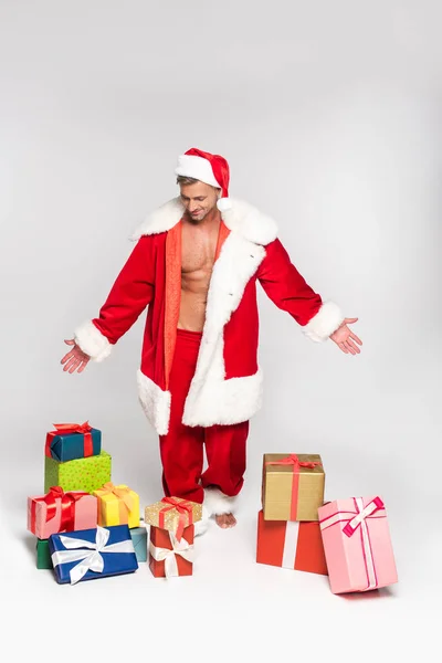 Schöner Mann im Weihnachtsmannkostüm schaut auf Geschenkschachteln auf grau — Stockfoto