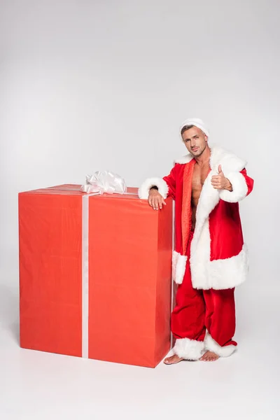Handsome santa claus leaning at big gift box and showing thumb up on grey — Stock Photo