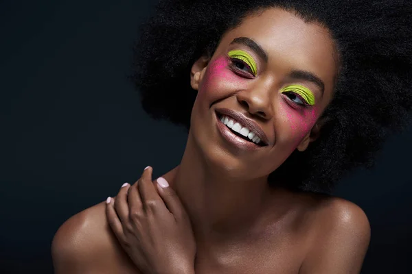 Retrato de modelo afroamericano sonriente con maquillaje de neón brillante posando aislado en negro - foto de stock
