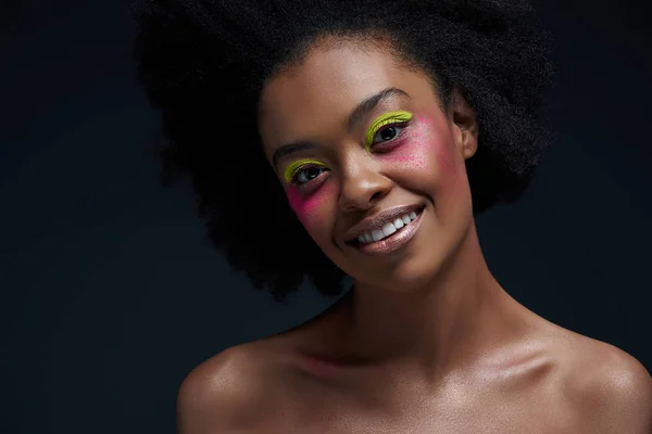 Retrato de modelo afroamericano sonriente con maquillaje de neón brillante posando aislado en negro - foto de stock