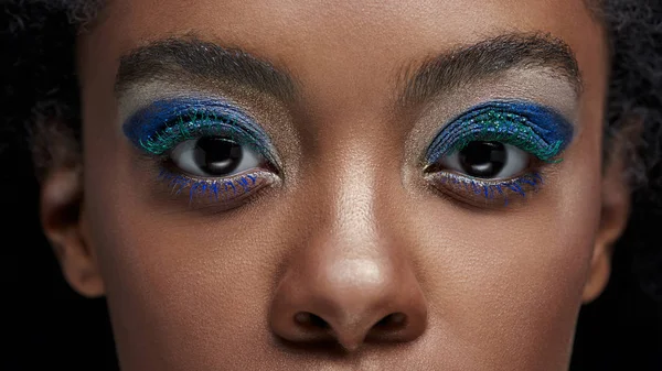 Plan recadré de femme afro-américaine avec des ombres à paupières bleues regardant la caméra isolée sur noir — Photo de stock