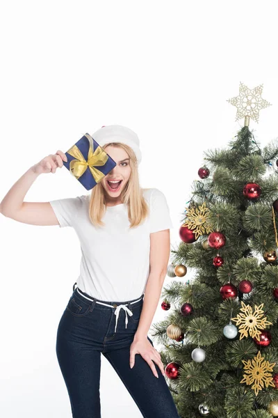 Збуджена жінка в капелюсі Санта Клауса з загорнутим подарунком біля ялинки ізольована на білому — стокове фото