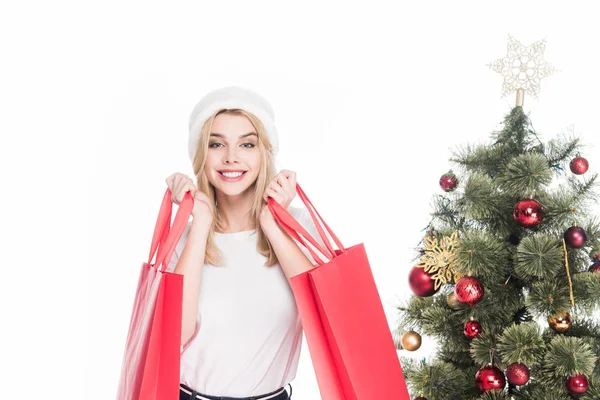 Portrait of smiling woman in santa claus hat with shopping bags near christmas tree isolated on white — Stock Photo
