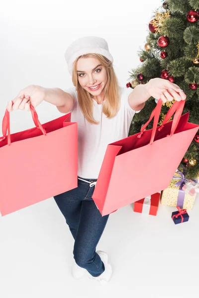 Vista de ángulo alto de la mujer sonriente en sombrero de Santa Claus con bolsas de compras cerca del árbol de Navidad aislado en blanco - foto de stock
