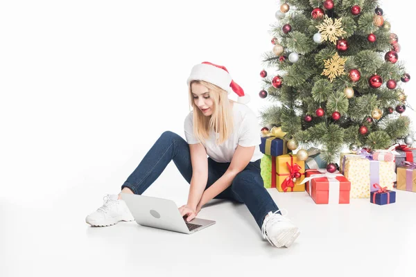 Mujer joven en sombrero de santa claus usando el ordenador portátil sentado cerca del árbol de Navidad sobre fondo blanco - foto de stock