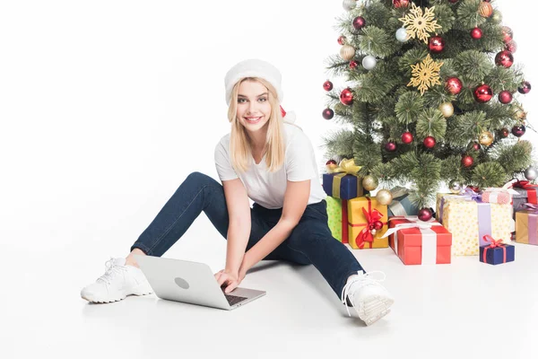 Усміхнена жінка в капелюсі Санта Клауса з ноутбуком, що сидить біля ялинки на білому тлі — стокове фото