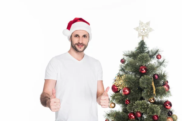 Portrait of young man in santa claus hat showing thumbs up near christmas tree isolated on white — Stock Photo