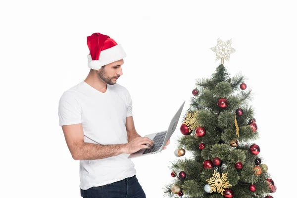 Homem em santa claus chapéu usando laptop perto de árvore de natal isolado em branco — Fotografia de Stock