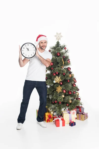 Hombre en sombrero de santa claus con el reloj de pie cerca del árbol de Navidad aislado en blanco - foto de stock