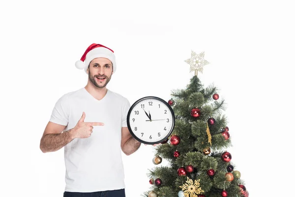 Retrato del hombre en sombrero de santa claus apuntando al reloj mientras está de pie cerca del árbol de Navidad aislado en blanco - foto de stock
