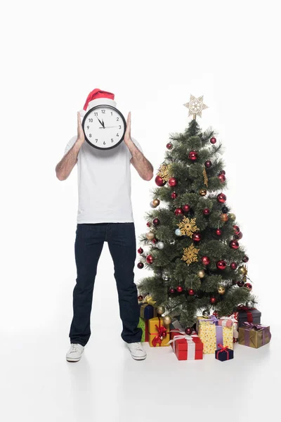 Vista oscura del hombre en sombrero de santa claus con el reloj de pie cerca del árbol de Navidad aislado en blanco - foto de stock