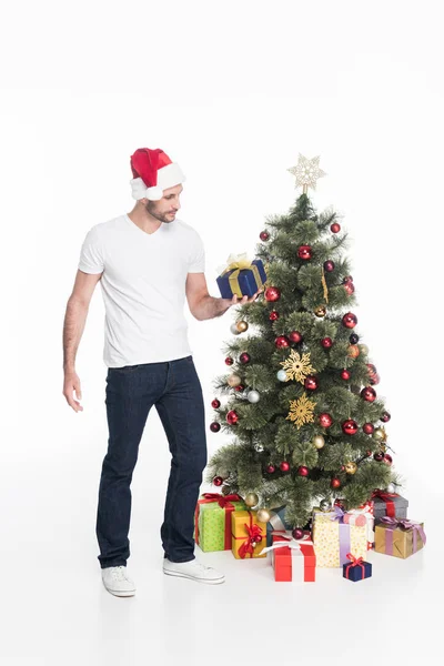 Joven en sombrero de santa claus con regalo envuelto de pie en el árbol de Navidad aislado en blanco - foto de stock