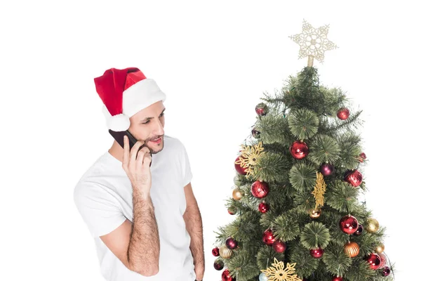 Человек в шляпе Санта-Клауса говорит на смартфоне возле рождественской елки, изолированной на белом — стоковое фото