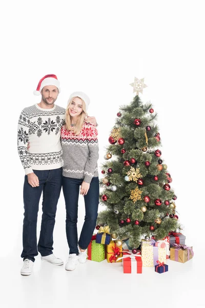 Casal sorridente em camisolas e chapéus de santa claus perto de árvore de natal isolado em branco — Fotografia de Stock