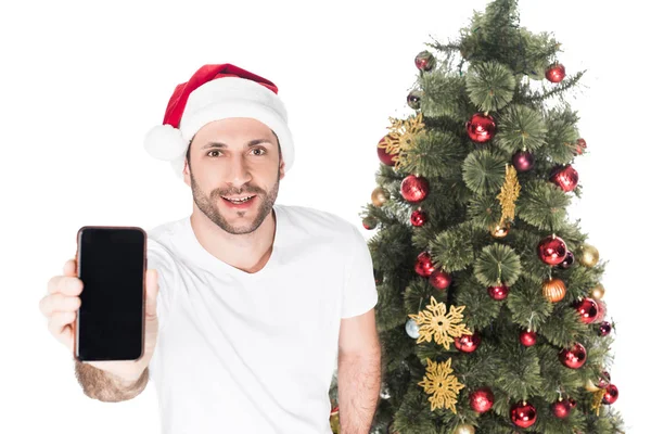 Hombre en sombrero de santa claus mostrando teléfono inteligente con pantalla en blanco cerca del árbol de Navidad aislado en blanco - foto de stock