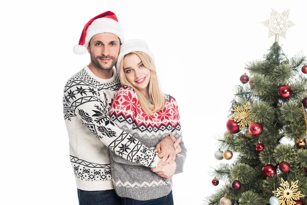 Joven pareja en santa claus sombreros abrazo cerca navidad árbol aislado en blanco - foto de stock