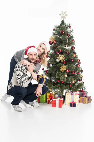 Couple in sweaters and santa claus hats hugging near christmas tree isolated on white — Stock Photo