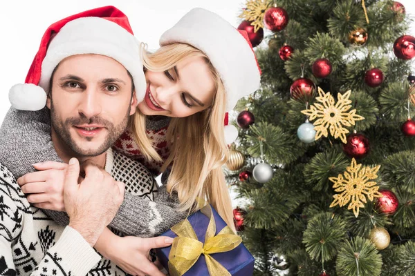 Retrato de pareja en sombreros de Santa Claus con regalo envuelto cerca del árbol de Navidad aislado en blanco - foto de stock