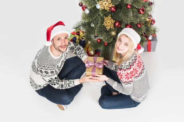 Alto ángulo vista de pareja en santa claus sombreros celebración de regalo envuelto cerca de árbol de Navidad aislado en blanco - foto de stock